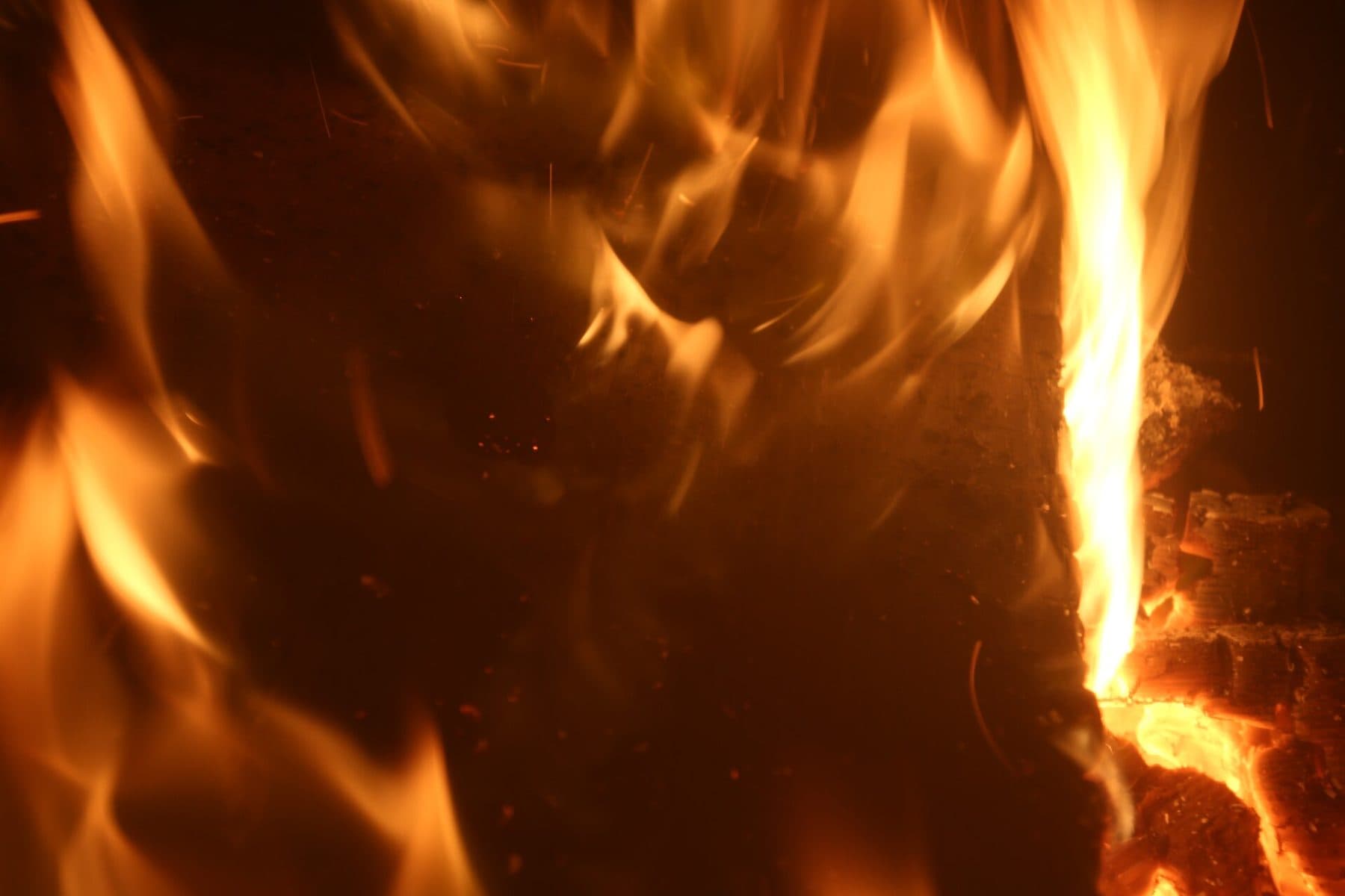 A hearth fire seen close up, tall gold and orange flames rising off glowing logs and embers against a dark surround.