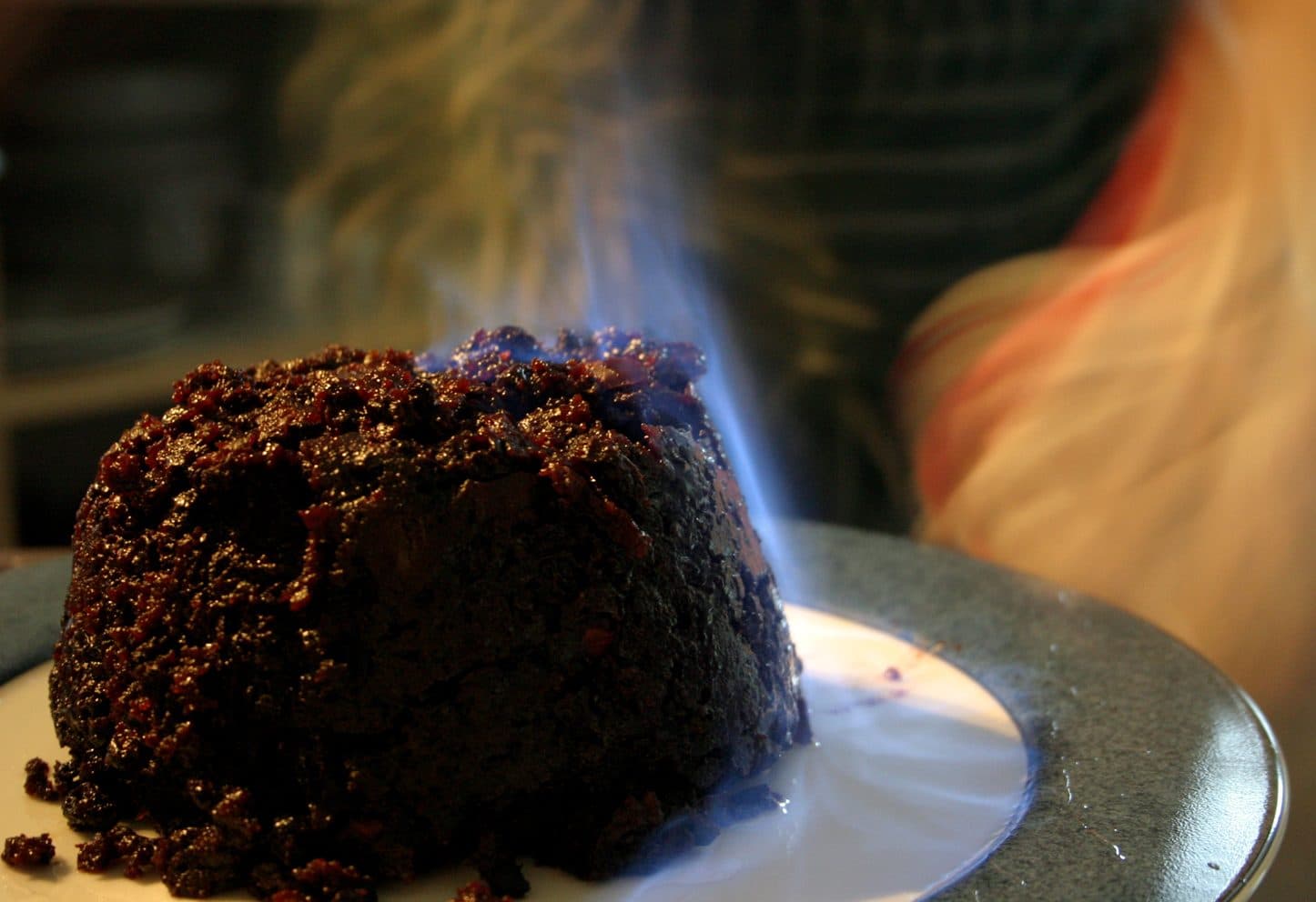 A dark, rich Christmas pudding on a plate with pale blue brandy flames flickering across the top, shot close up in a warm, dim room.