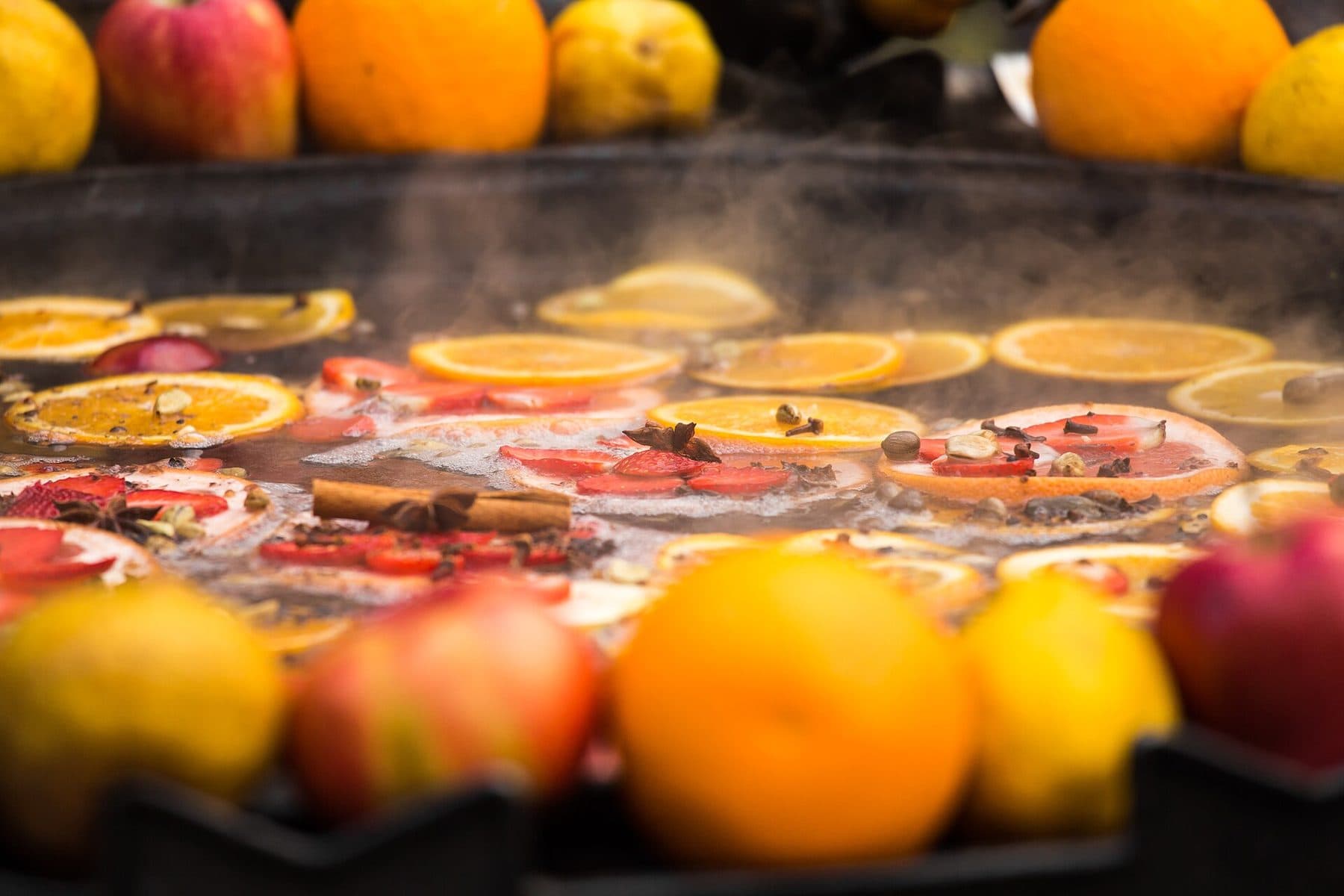 A wide steaming pot of mulled wine studded with floating orange and lemon slices, cinnamon sticks and star anise, ringed by whole oranges and apples, steam rising off the surface.
