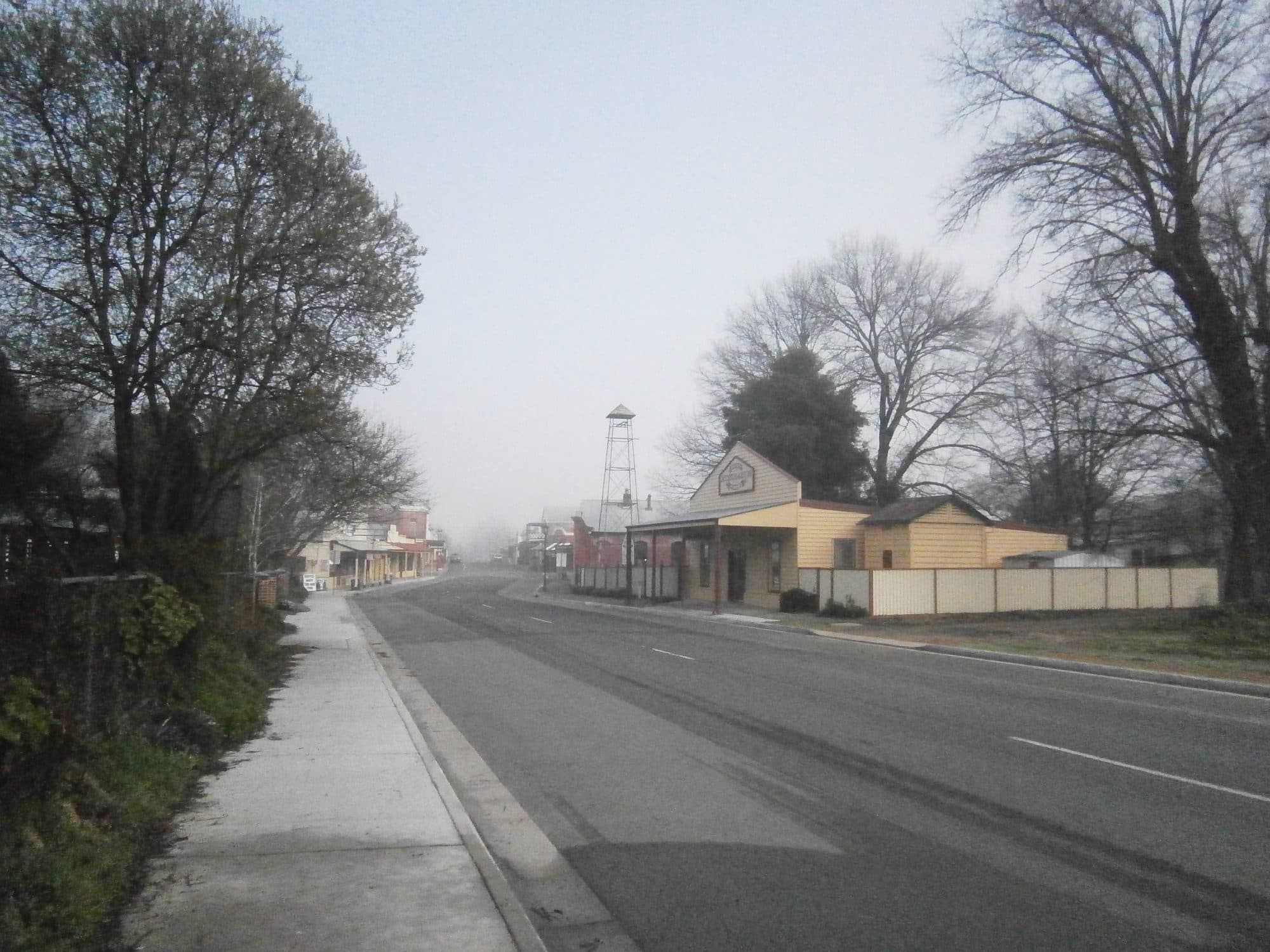 Conness Street in Chiltern, Victoria, looking west — wide road, low historic shopfronts on both sides, mature trees, a quiet autumn morning.