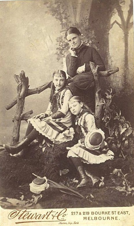 A formal Victorian-era studio portrait of a mother standing behind two young girls seated on a rustic wooden fence prop, all in dark high-collared dress, against a painted woodland backdrop.