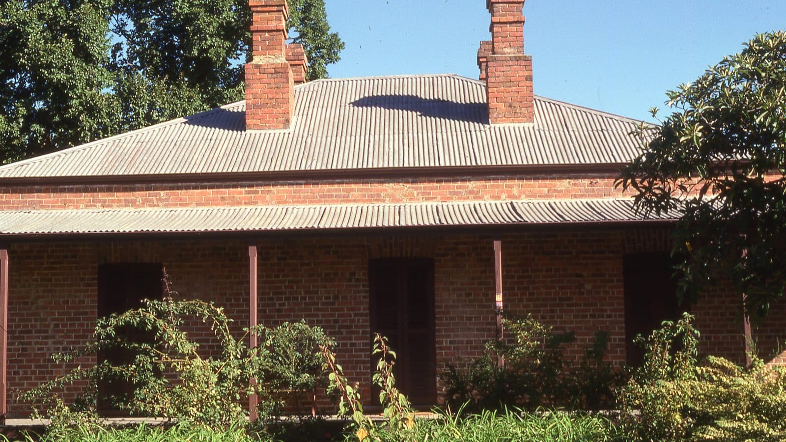 A single-storey red brick house with a corrugated iron roof, two tall chimneys and a front verandah, with garden plants in the foreground and a blue sky behind.