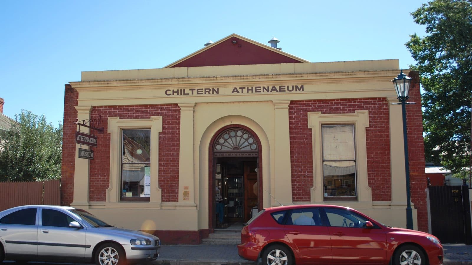 The cream and red brick facade of the Chiltern Athenaeum, a small single storey heritage building with an arched doorway and a raised pediment reading Chiltern Athenaeum, on a quiet street.