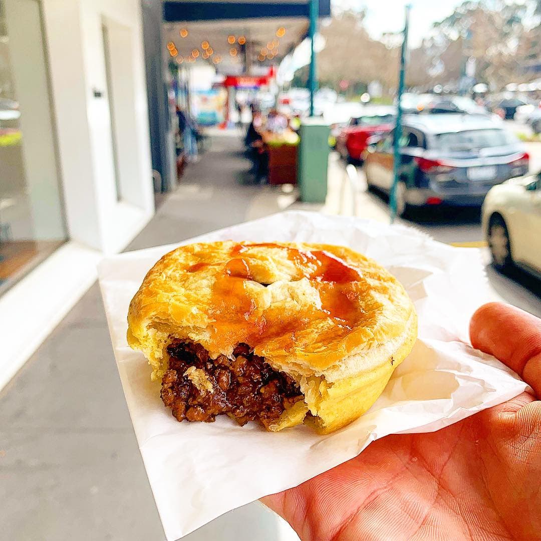 A hand holds a golden Australian meat pie with a bite taken out, the minced-beef filling and a squiggle of tomato sauce showing, a country street behind.