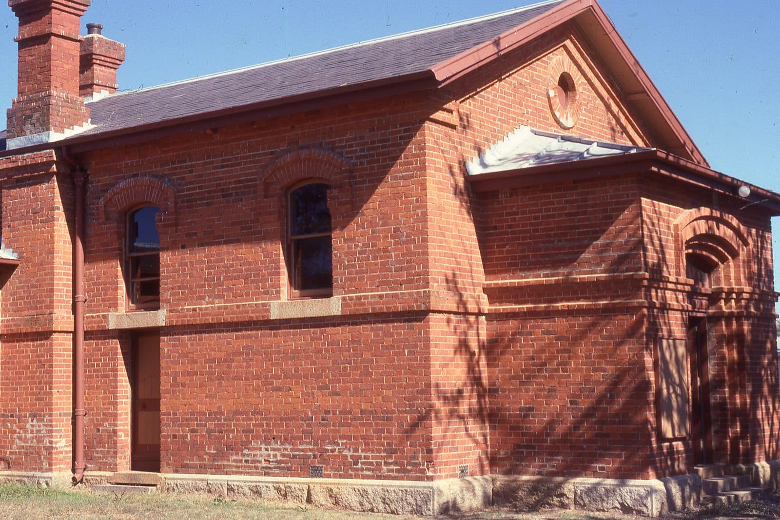 A small nineteenth century red brick courthouse with tall arched windows, brick chimneys and a stone base, photographed from the corner in bright sun.