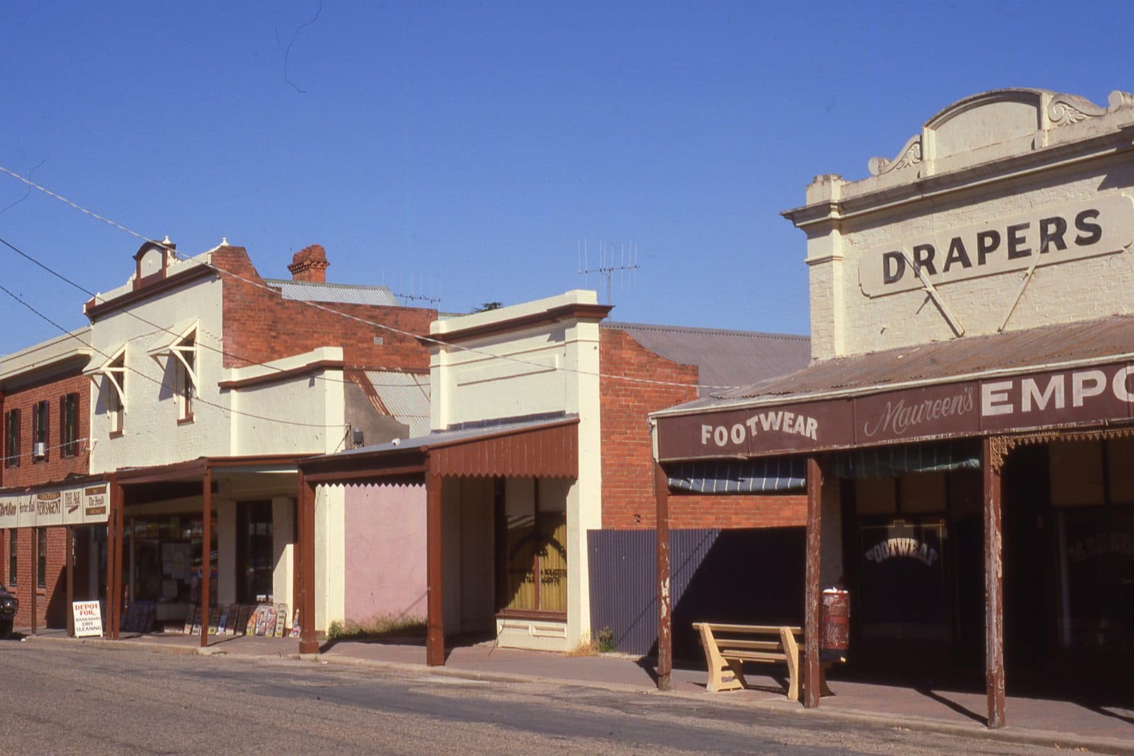 Old shopfronts on a country main street, one with DRAPERS across the parapet above a footwear and emporium verandah, timber posts holding up the awning and a wooden bench on the footpath.