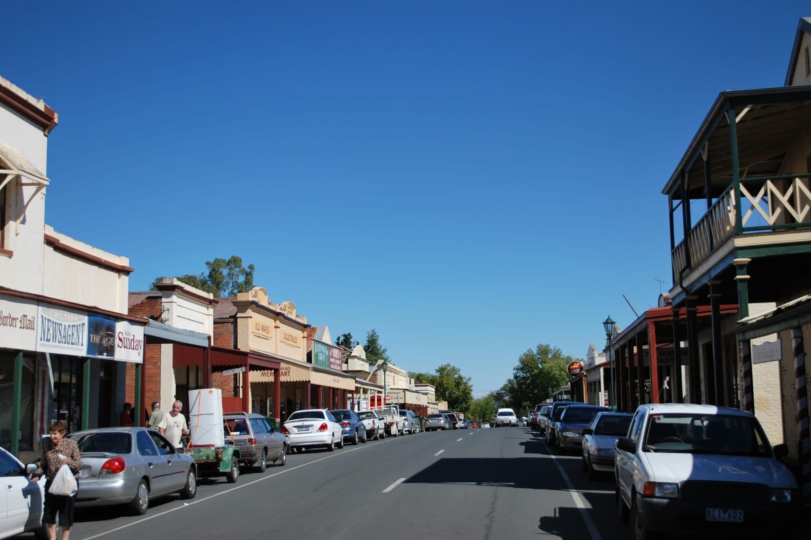Chiltern's main street in the present day, cars parked at an angle along both kerbs, people on the footpath, and the same verandahs and painted shopfronts running away down the road.