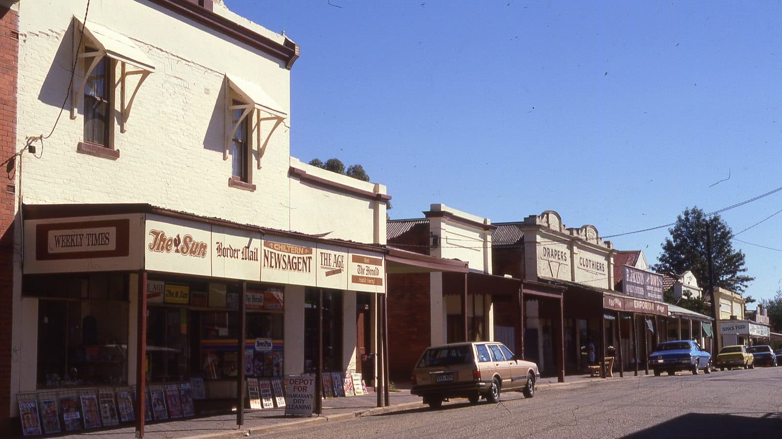 A row of nineteenth century shopfronts along a country main street, a newsagent with painted newspaper signs across its verandah, and drapers and emporium signage further down the row under a clear blue sky.